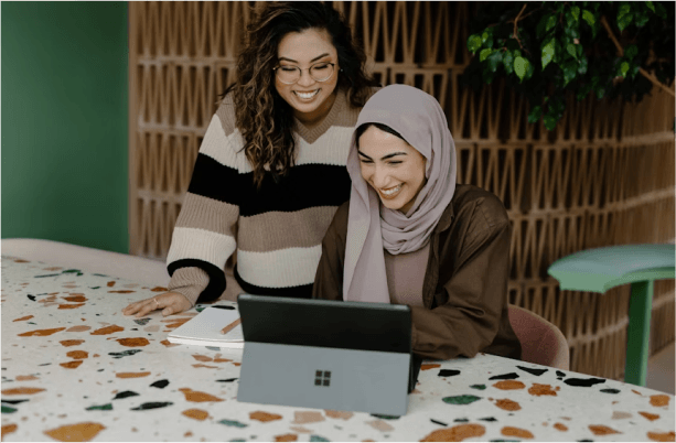 Two smiling women collaborate on a Microsoft Surface laptop, reflecting teamwork and innovation—core values of a results-driven digital marketing agency.